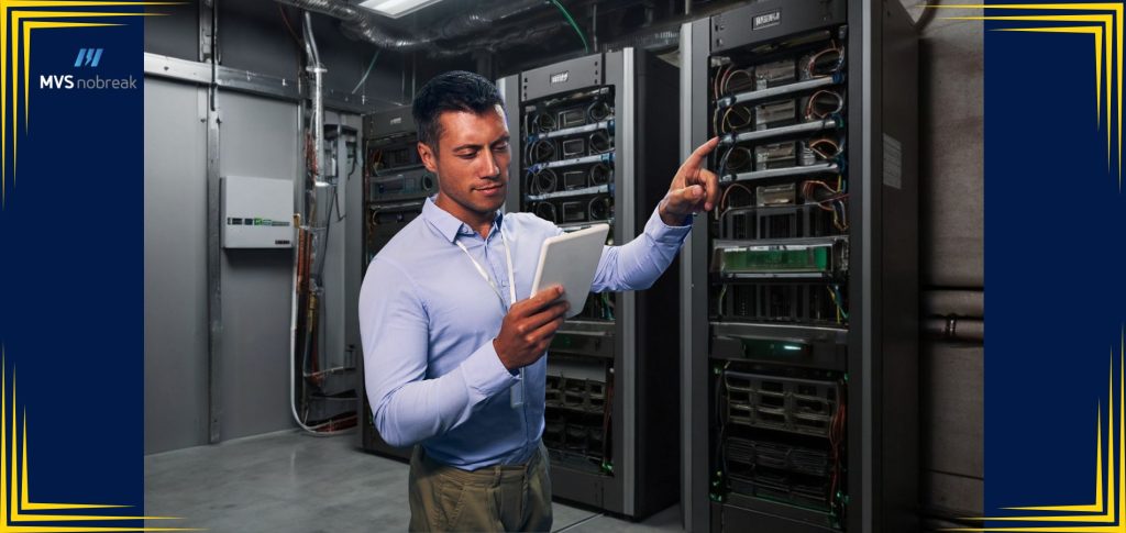 Na imagem, um técnico está realizando a manutenção em um ambiente de servidor com vários nobreaks. Ele está usando um tablet para verificar informações e parâmetros, enquanto aponta para um dos equipamentos. A imagem reflete a importância da manutenção preventiva e da monitoração constante dos nobreaks para garantir a continuidade e a proteção dos sistemas críticos. Este é o cenário ideal para ilustrar o tema do artigo "Guia Completo de Manutenção de Nobreaks", destacando as práticas recomendadas e os cuidados necessários para manter esses equipamentos em perfeito funcionamento.