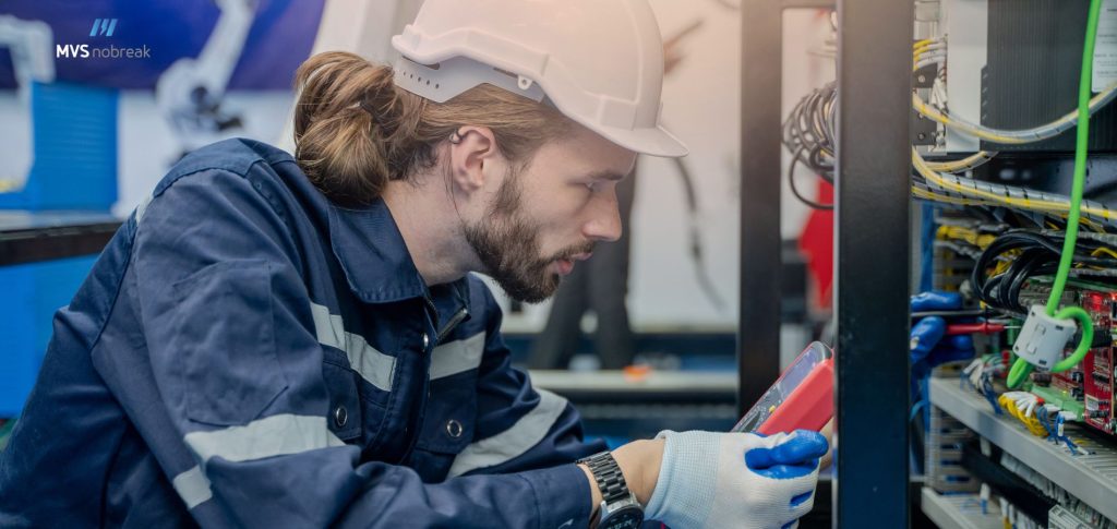 Na imagem, um técnico de manutenção está inspecionando um nobreak, utilizando um equipamento de medição. Ele veste um uniforme de segurança com capacete e luvas, refletindo o compromisso com a segurança no trabalho. Este cenário enfatiza a importância da manutenção e cuidado com nobreak para garantir a operação contínua e eficiente dos sistemas elétricos.