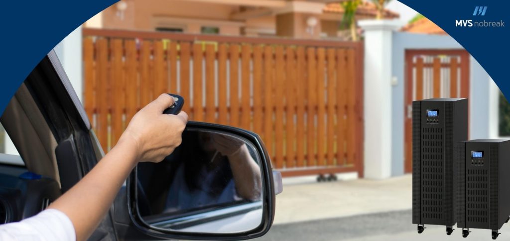 **Descrição da Imagem:** A imagem mostra uma pessoa dentro de um carro usando um controle remoto para abrir um portão eletrônico de madeira. No canto direito, aparecem dois nobreaks de porte médio, simbolizando a importância desses dispositivos para o funcionamento do portão em caso de falhas de energia. A cena transmite segurança e comodidade, destacando o papel do nobreak na operação contínua e protegida do portão eletrônico. No canto superior direito, há o logotipo da MVS Nobreak, reforçando a marca.